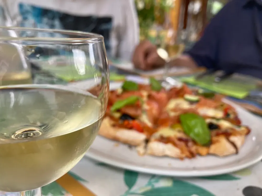 Pizza garnie de légumes et basilic, servie en terrasse avec un verre de vin blanc